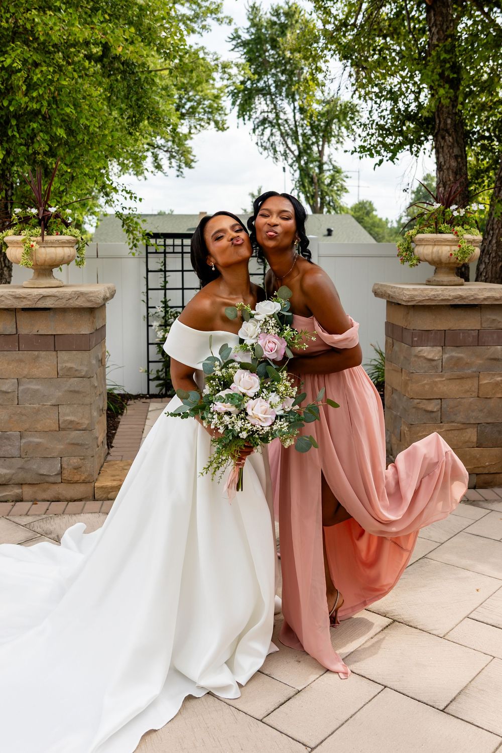 Playful wedding photo with bride and bridesmaid posing dramatically on outdoor stone patio.