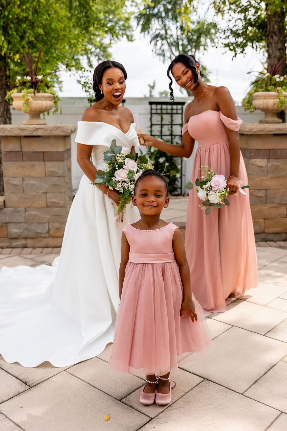 Bride and bridesmaid pose with flower girl in matching pink dress on stone patio.