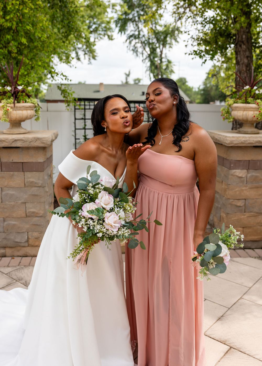 Bride and bridesmaid share fun moment while posing with bouquet outdoors.
