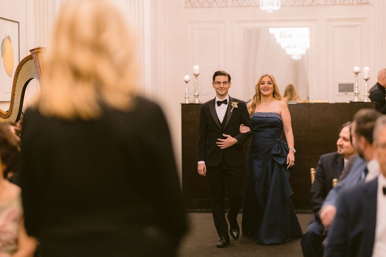 Groom walking down the aisle in formal attire enter a grand ballroom with elegant white decor and chandeliers.