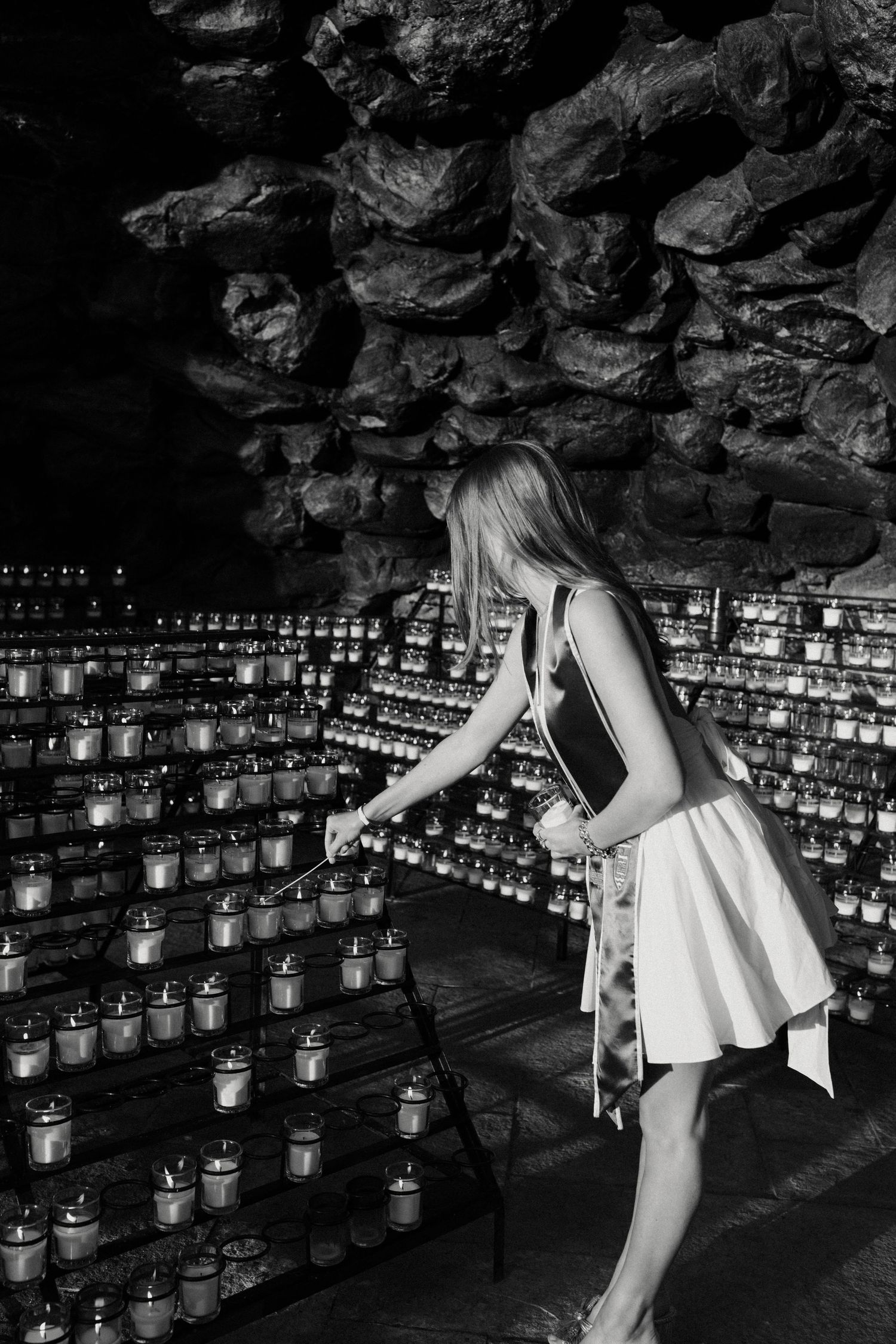 Person in white dress lighting candles in dark grotto with stone walls.