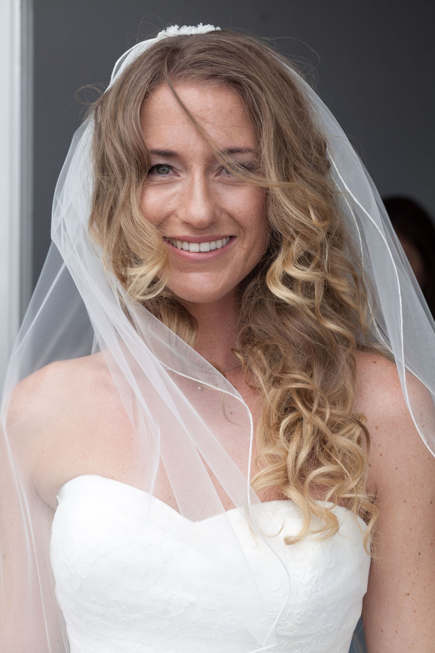 Bride wearing white wedding dress with long curled blonde hair and delicate veil smiling at camera.