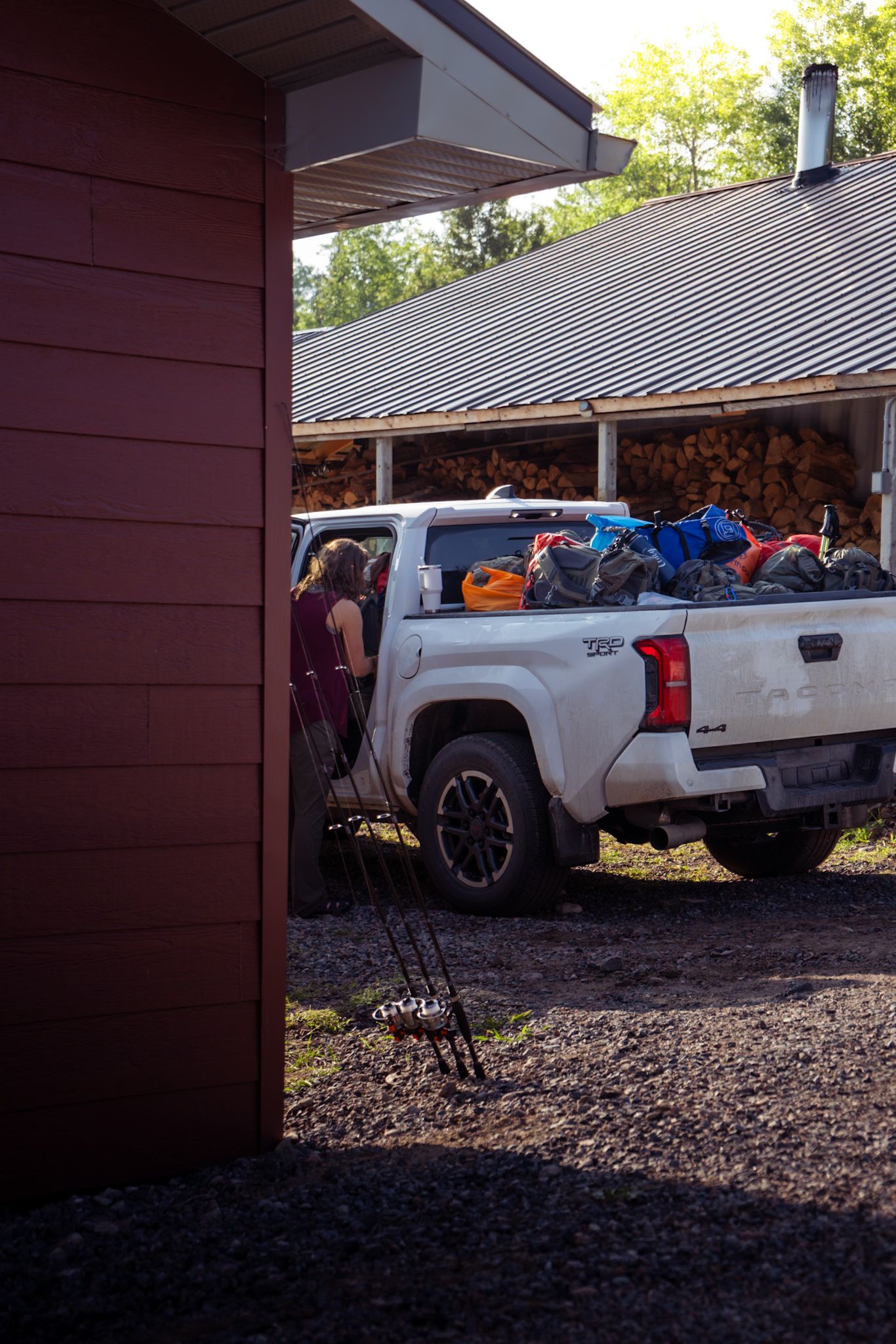 A modern Chevy Silverado pickup truck loaded with gear parked under a covered storage area with stacked firewood.