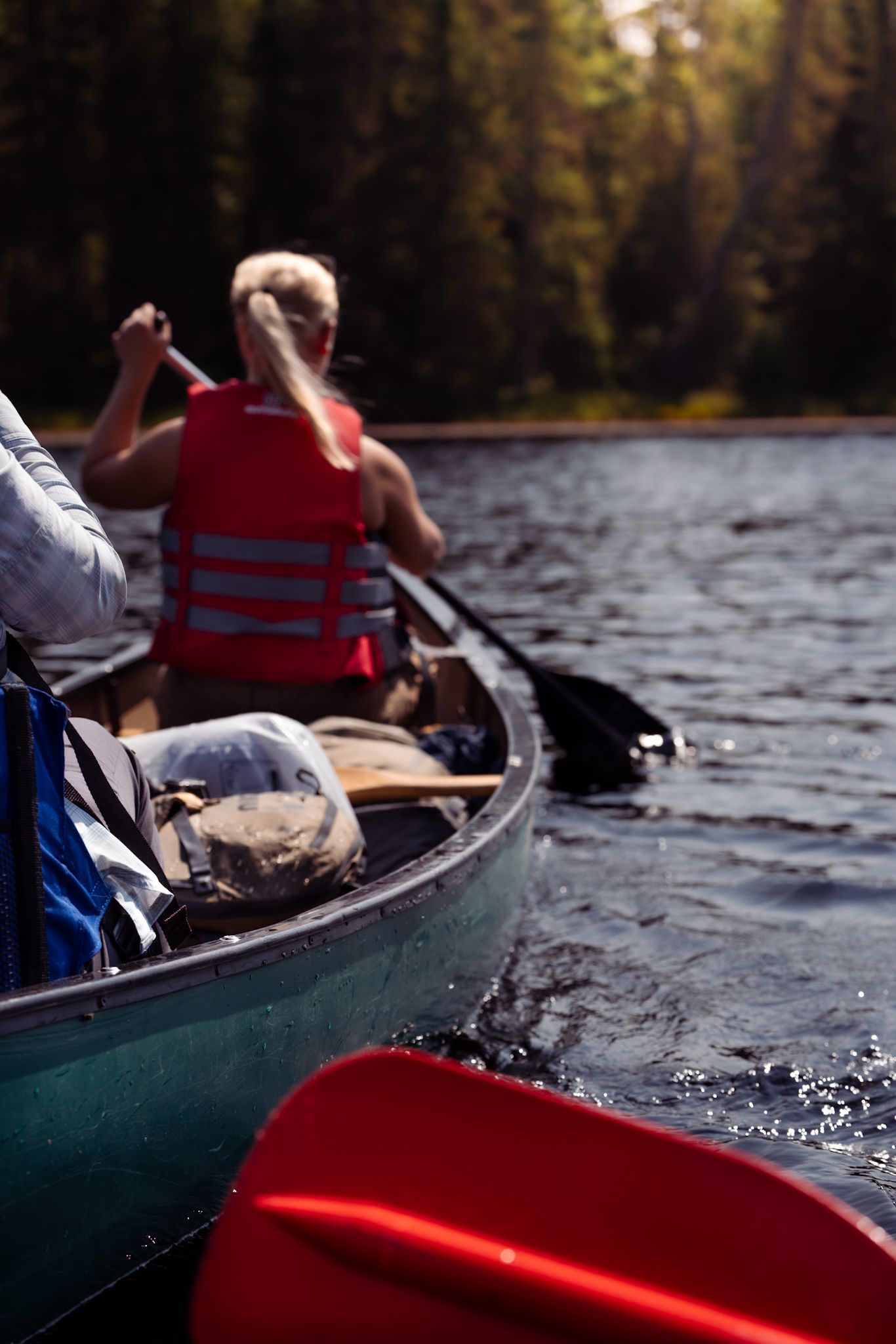 Someone paddling a green canoe on a calm lake while wearing a red life vest, with gear stowed in the vessel.