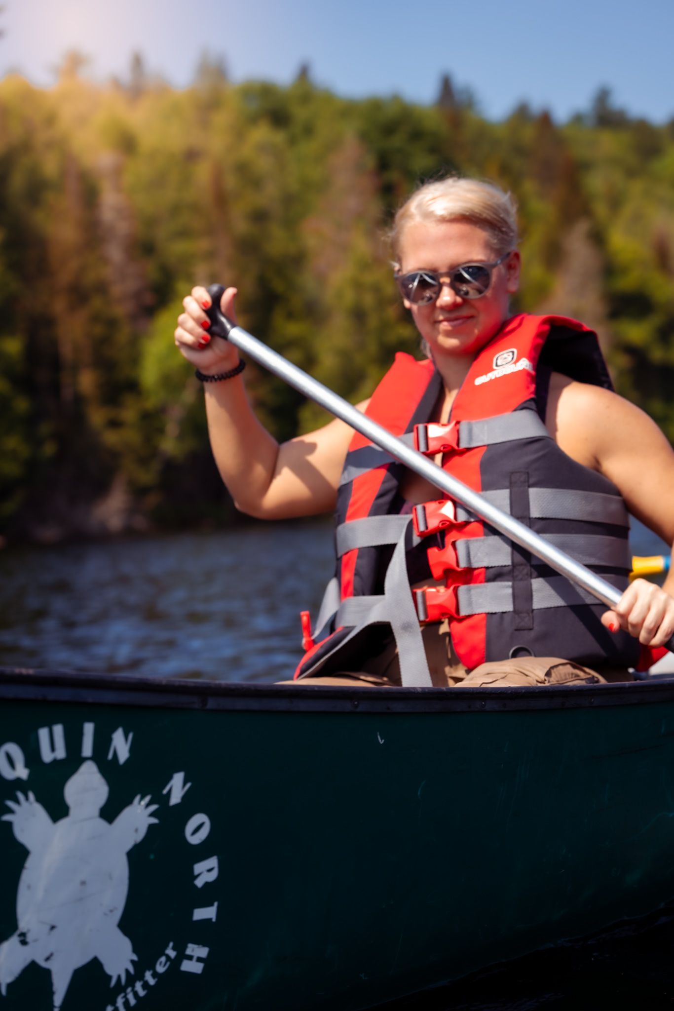Person in red life vest paddling a dark green canoe on a lake with forest in the background on a sunny day.
