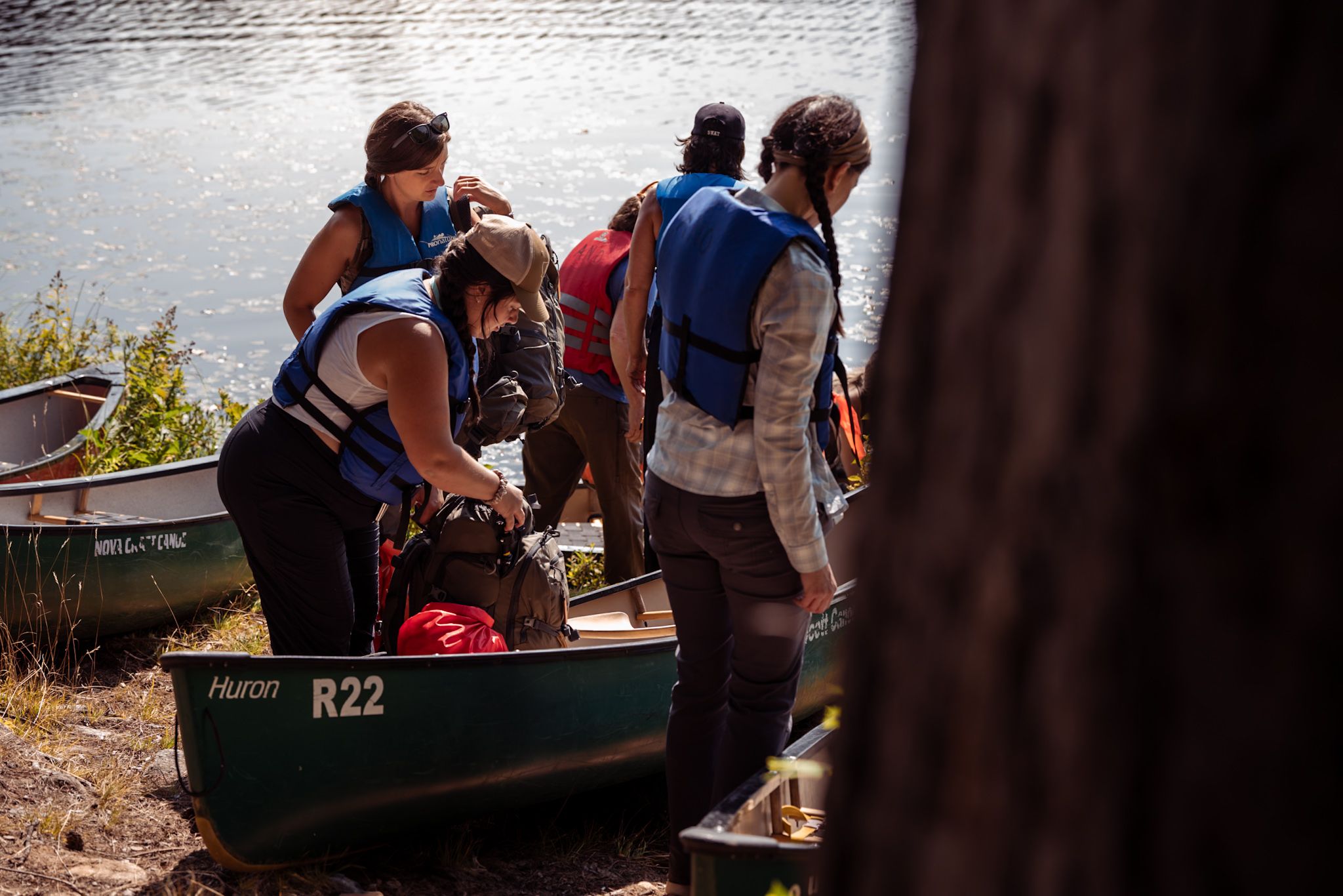 A group of people preparing to embark on a canoeing adventure, loading gear into a green R22 canoe by the water.