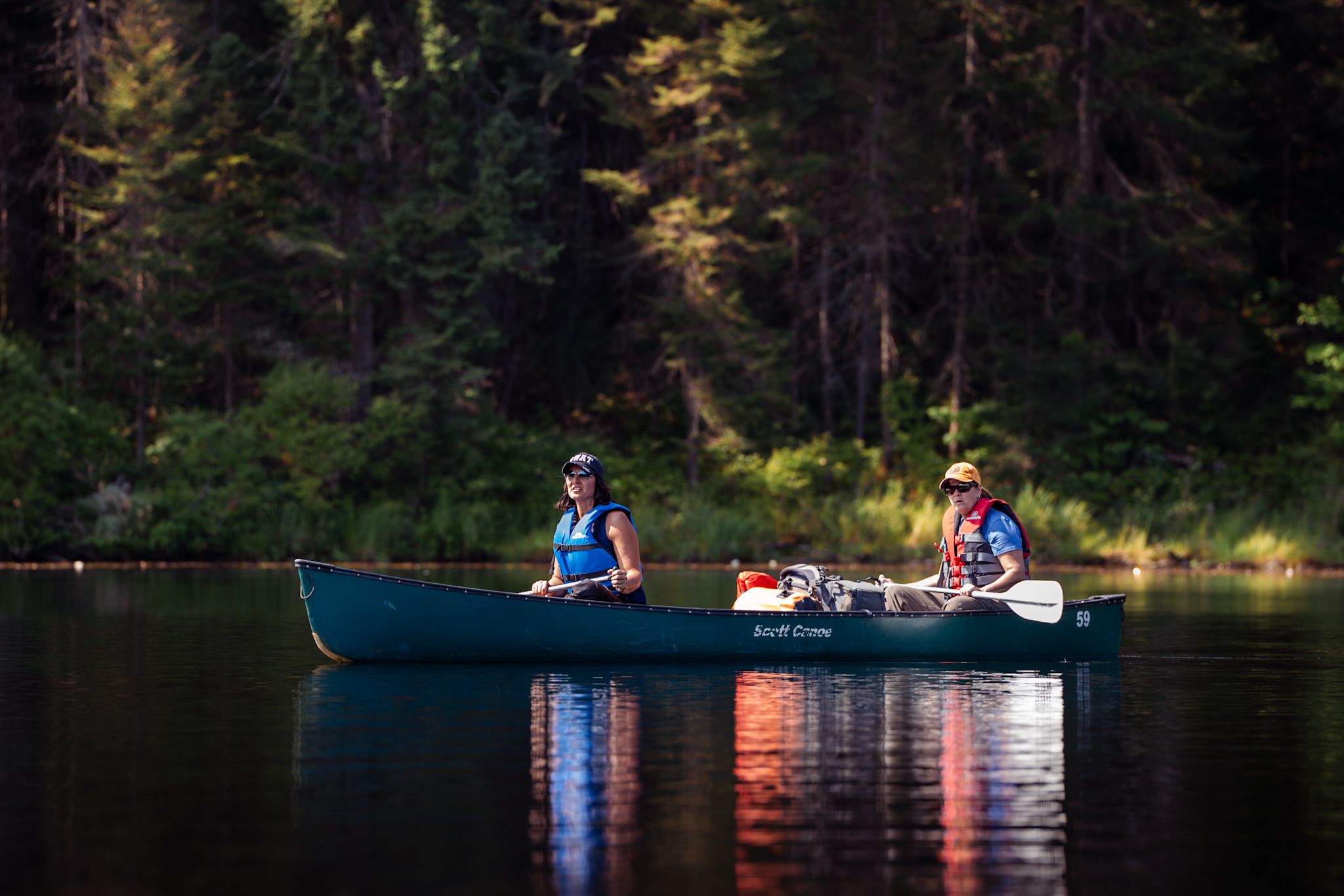 Two people paddle a blue canoe across a calm lake with sunlit forest reflections in the background.