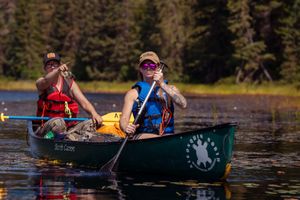 Two people paddling a green canoe across a lake with evergreen trees along the shoreline in summer.