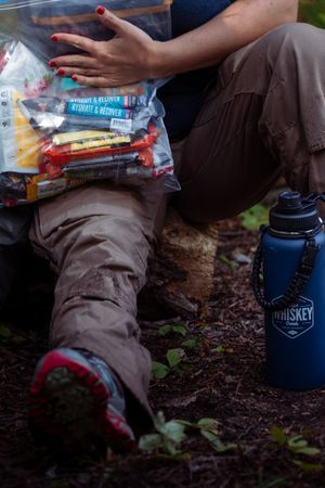 A hiker rests with snacks and a blue Whiskey Peaks water bottle on a dirt trail in the woods.