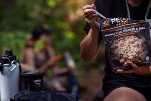 A close-up shot of someone holding a Peak frozen pizza while camping with outdoor gear visible in the background.