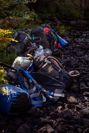 Multiple hiking backpacks and camping gear scattered on rocky ground near a stream in low evening light.
