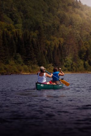 Two people paddle a green canoe across a serene lake surrounded by dense forest on a summer day.