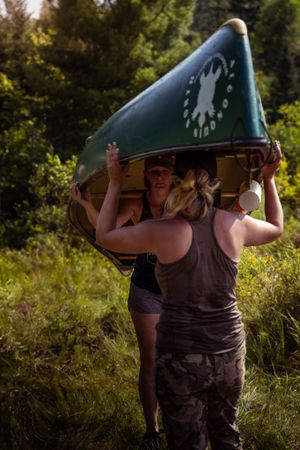 Two people carrying a green canoe overhead on a sunny summer day in a wooded area.