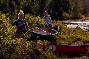 Two people prepare red canoes for an outdoor adventure along a scenic riverside surrounded by lush vegetation.