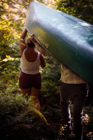 Two people carrying a green canoe through a forest trail on a sunny day.