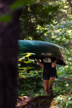 A kayaker carrying their boat along a forest trail wears a North Face shirt and shorts amidst lush greenery.