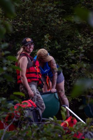 Two people in life jackets prepare to launch a canoe into water through leafy foliage.