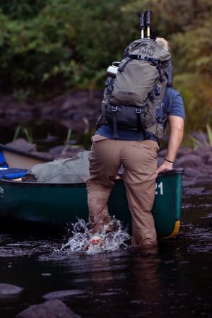 A hiker with backpack wades through shallow river water next to a green canoe in a rocky stream.