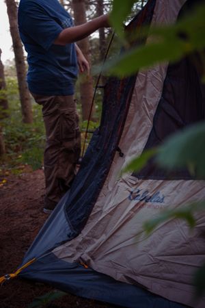 Close-up of someone setting up a beige and navy camping tent in a wooded forest setting at dusk.