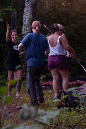 Three people gather with camping gear in a wooded forest area at dusk.
