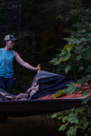 Person in blue tank top and cap adjusts gear in a canoe surrounded by leafy forest foliage at dusk.
