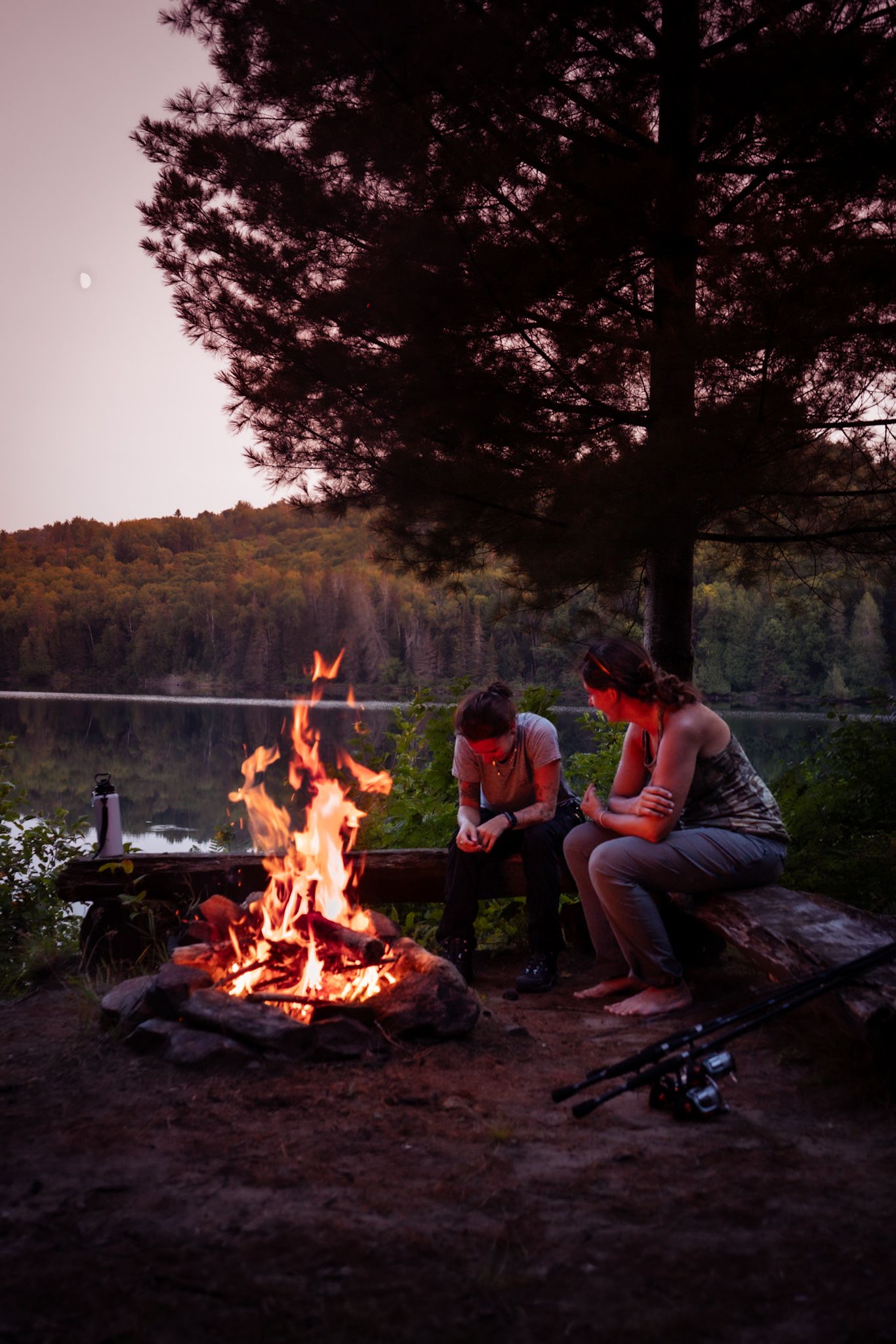 People gathered around a campfire at dusk by a lake, with trees and hills silhouetted in the background.
