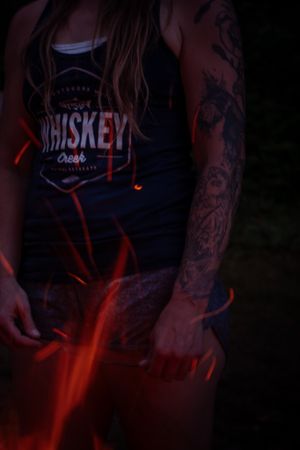A person wearing a black Whiskey tank top with visible arm tattoos illuminated by red lighting in a dark setting.