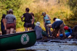 A green canoe sits by the shoreline while a group of people gather near the water's edge on a sunny day.