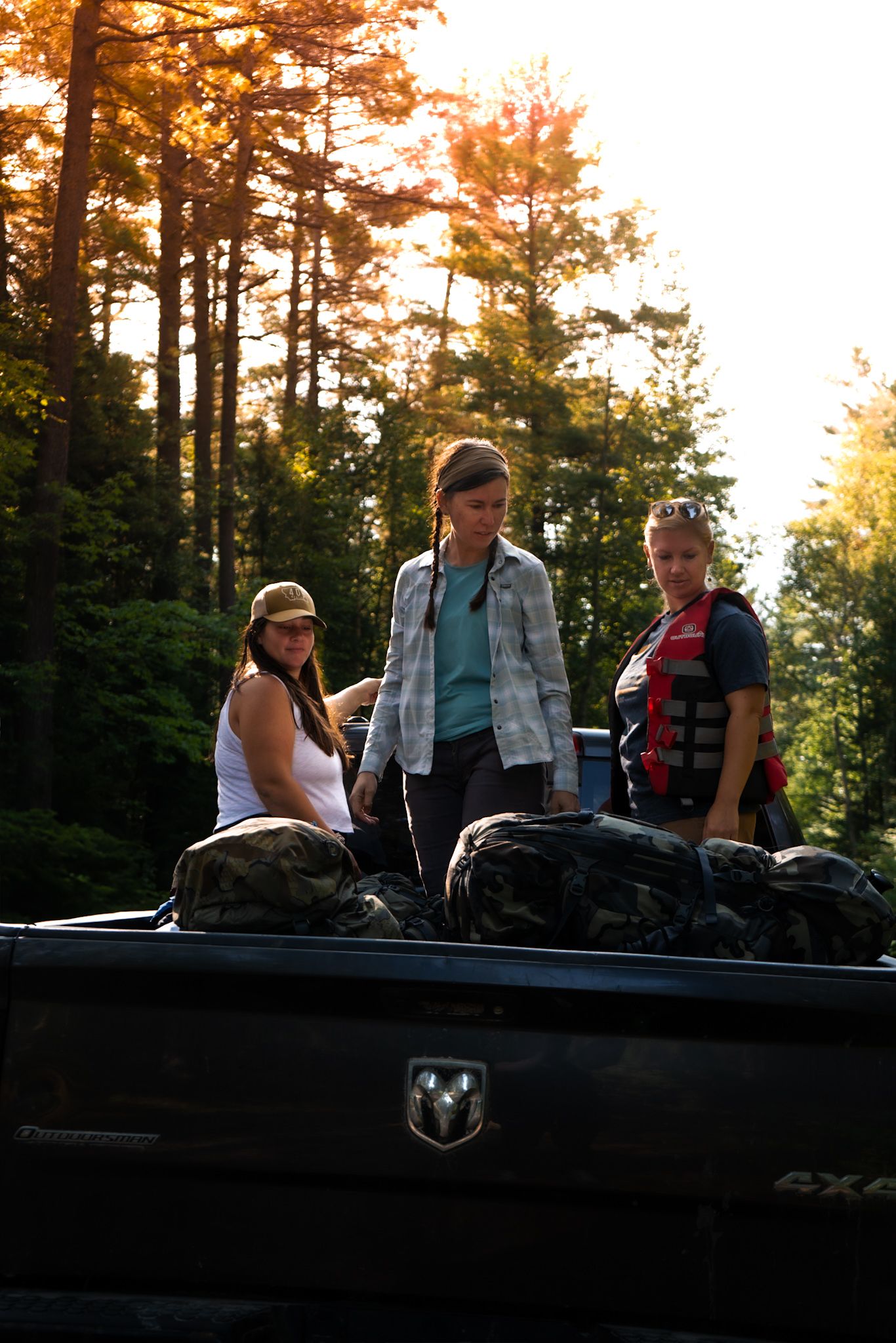 Three people stand in the back of a pickup truck in a sunlit forest during golden hour.