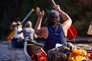 Paddler in a blue tank top navigates a canoe loaded with gear and supplies down a river on a sunny day.
