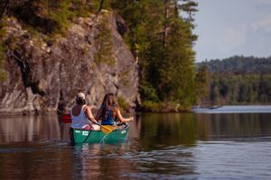 Two people paddle a green canoe along calm waters near rocky cliffs and evergreen trees on a scenic lake.