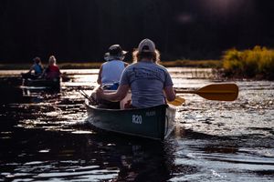 People paddling canoes on a tranquil lake at sunset with golden light reflecting on the rippling water.