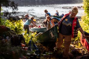 Group of adventurers work together to carry a canoe up a sunlit slope near a river on a summer day.