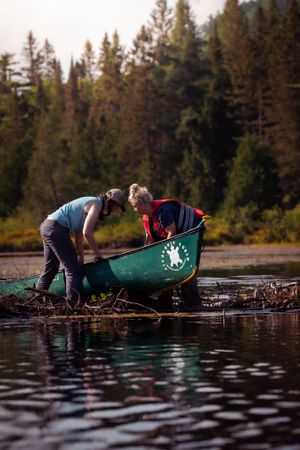 Two people in a green canoe explore a peaceful lake surrounded by evergreen forest at dusk.