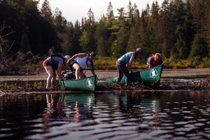 People gather cranberries from canoes on a dark reflective lake surrounded by evergreen forest at dusk.
