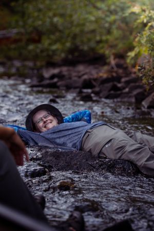 Person relaxing and laying in a shallow rocky stream while wearing a hat and blue shirt.