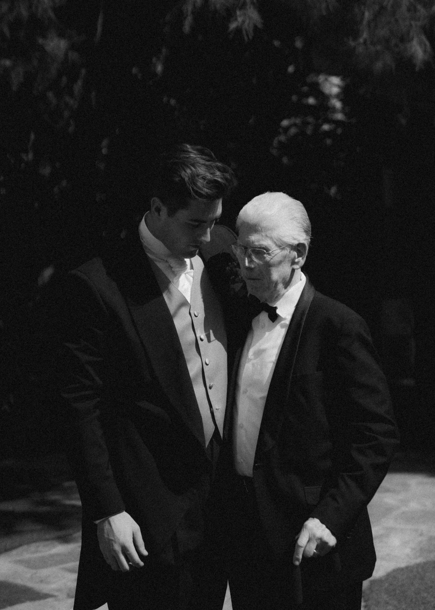 Two elegantly dressed men in black tuxedos engaged in conversation at a formal event in black and white photography.