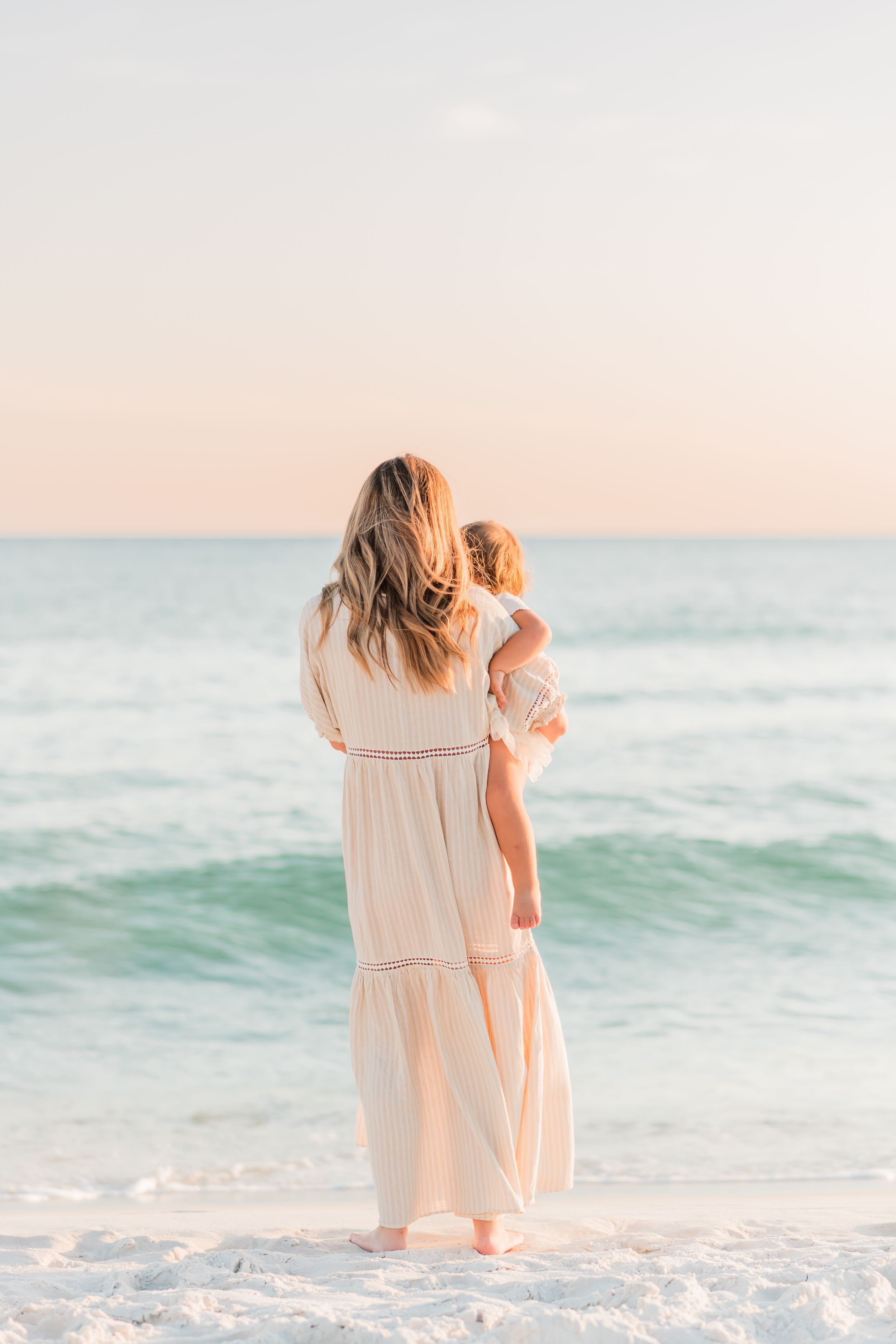 A person in a flowing white dress holds a child while standing on a peaceful beach at sunset.