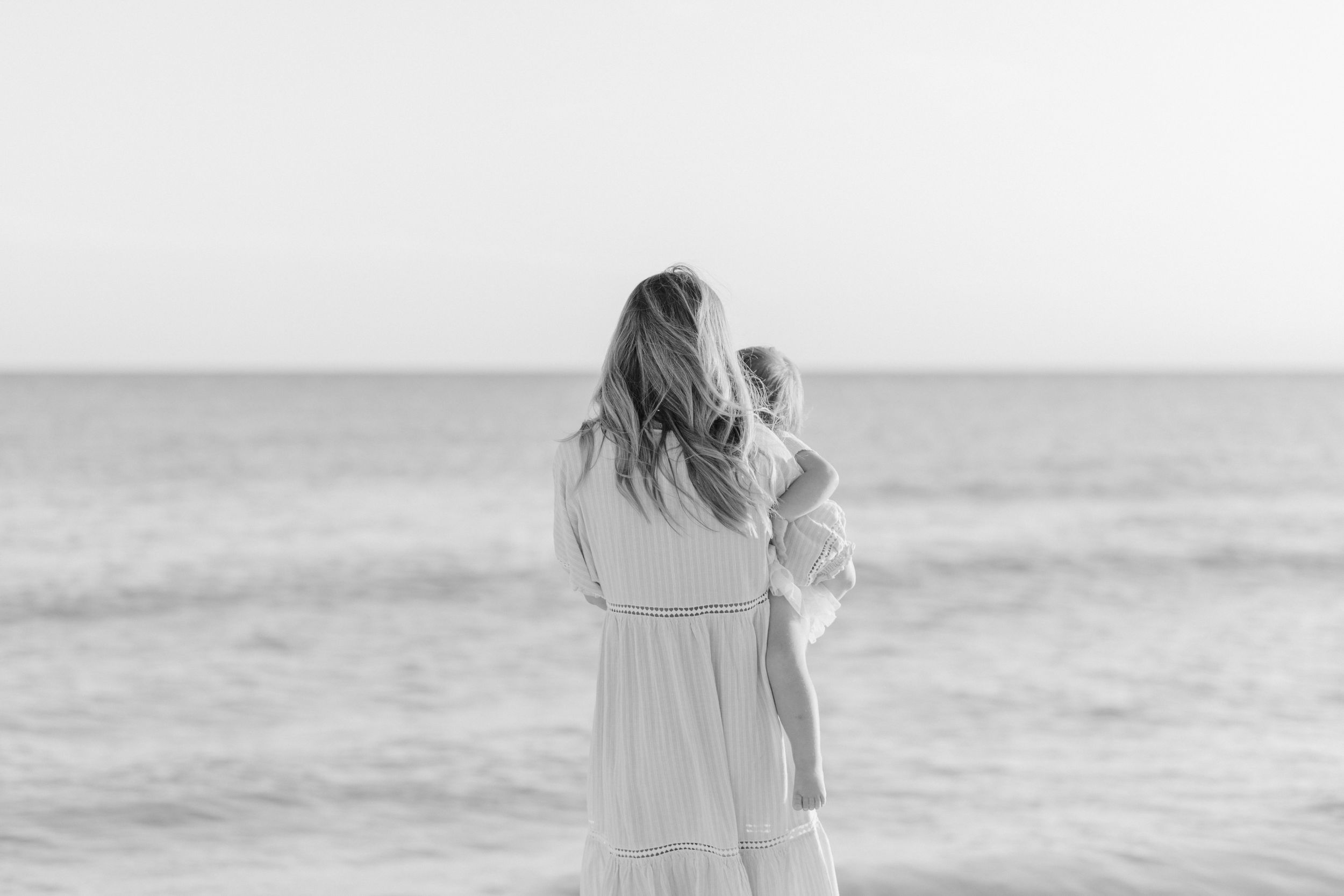 Black and white photo of someone in a flowing dress standing at the ocean's edge looking out at the horizon.