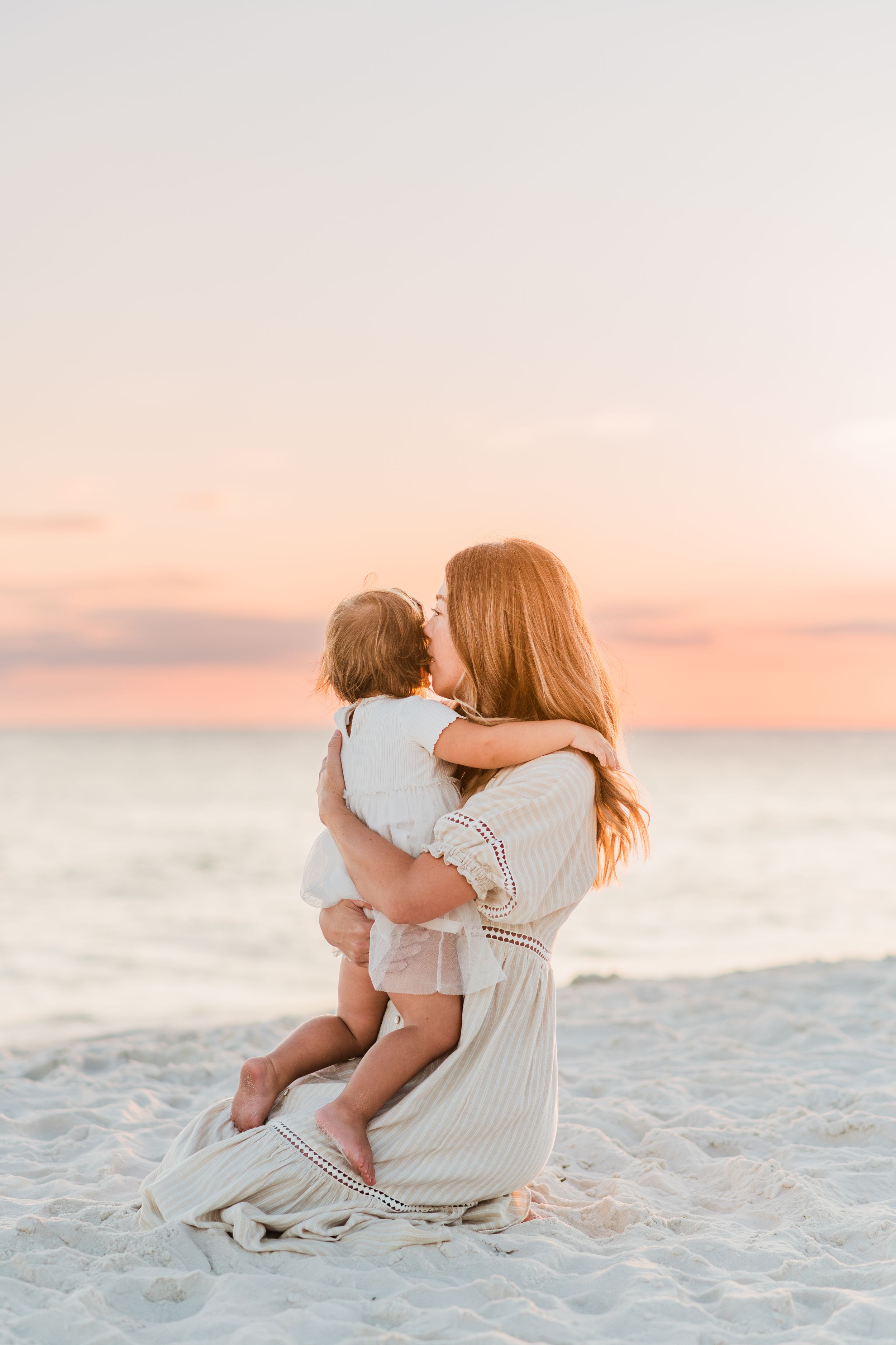A tender beach scene at sunset with figures in white dresses sitting together on pristine sand by the ocean.