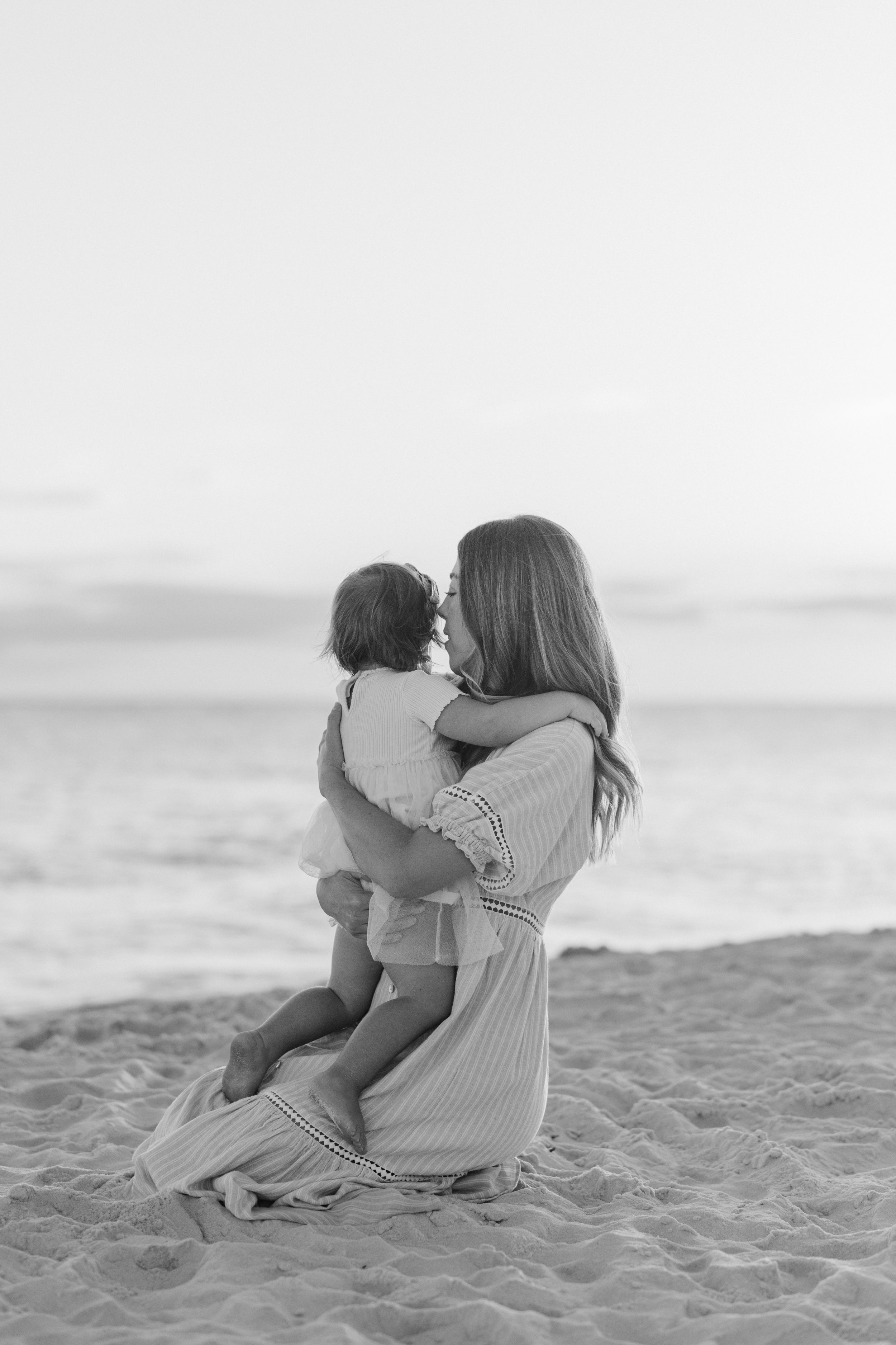 A touching black and white moment between parent and child sitting together on a sandy beach at sunset.