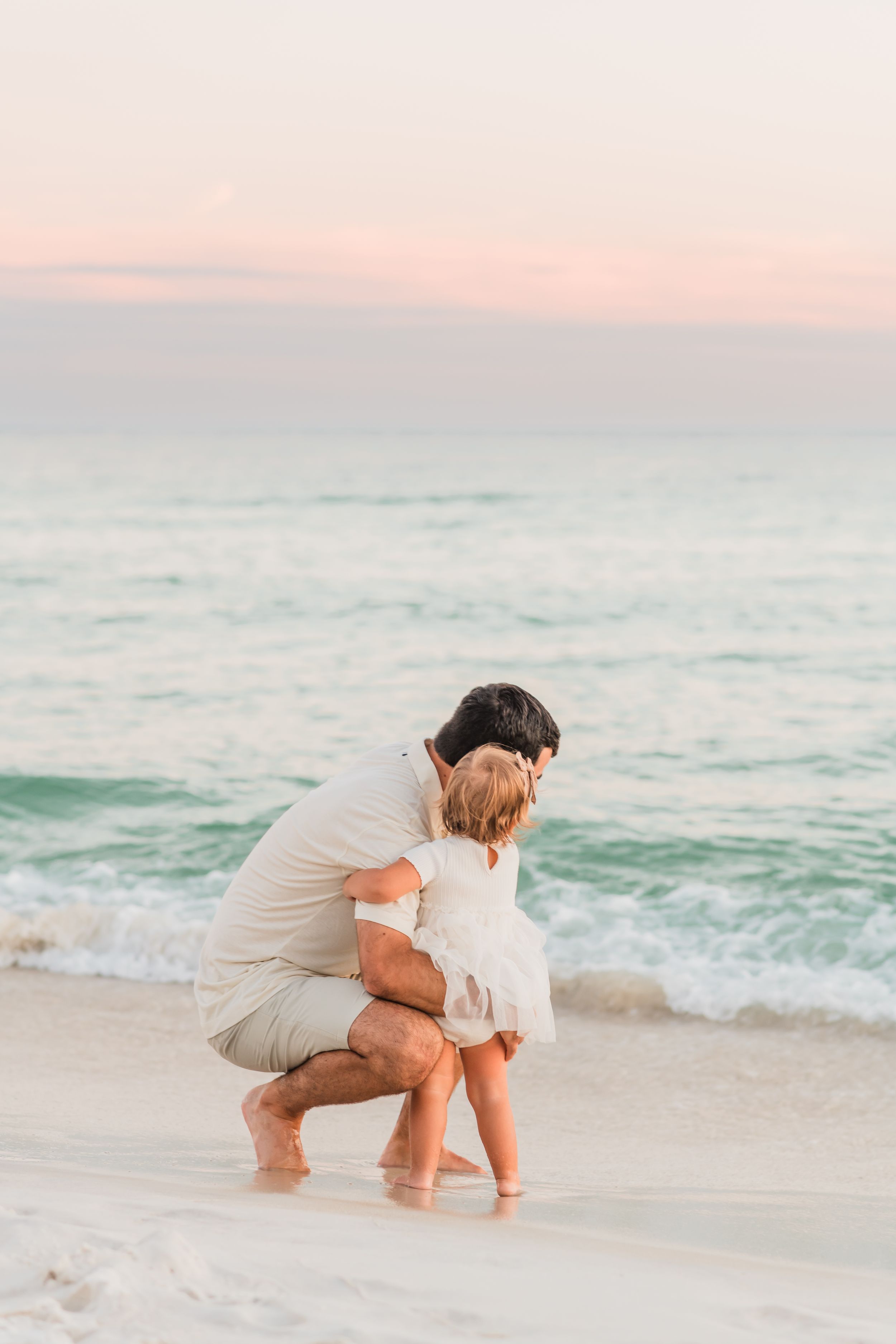 An adult and child share a tender moment at the beach during sunset while standing in the surf.