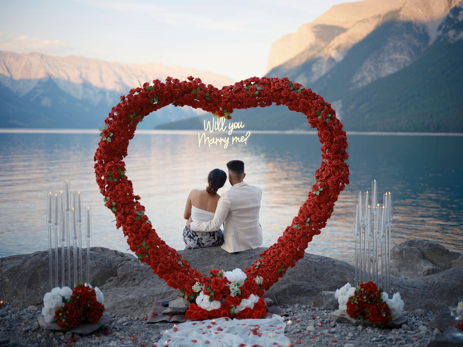A couple sits beneath red heart-shaped floral arches overlooking a tranquil lake and mountain landscape at sunset in Banff in the Rockies