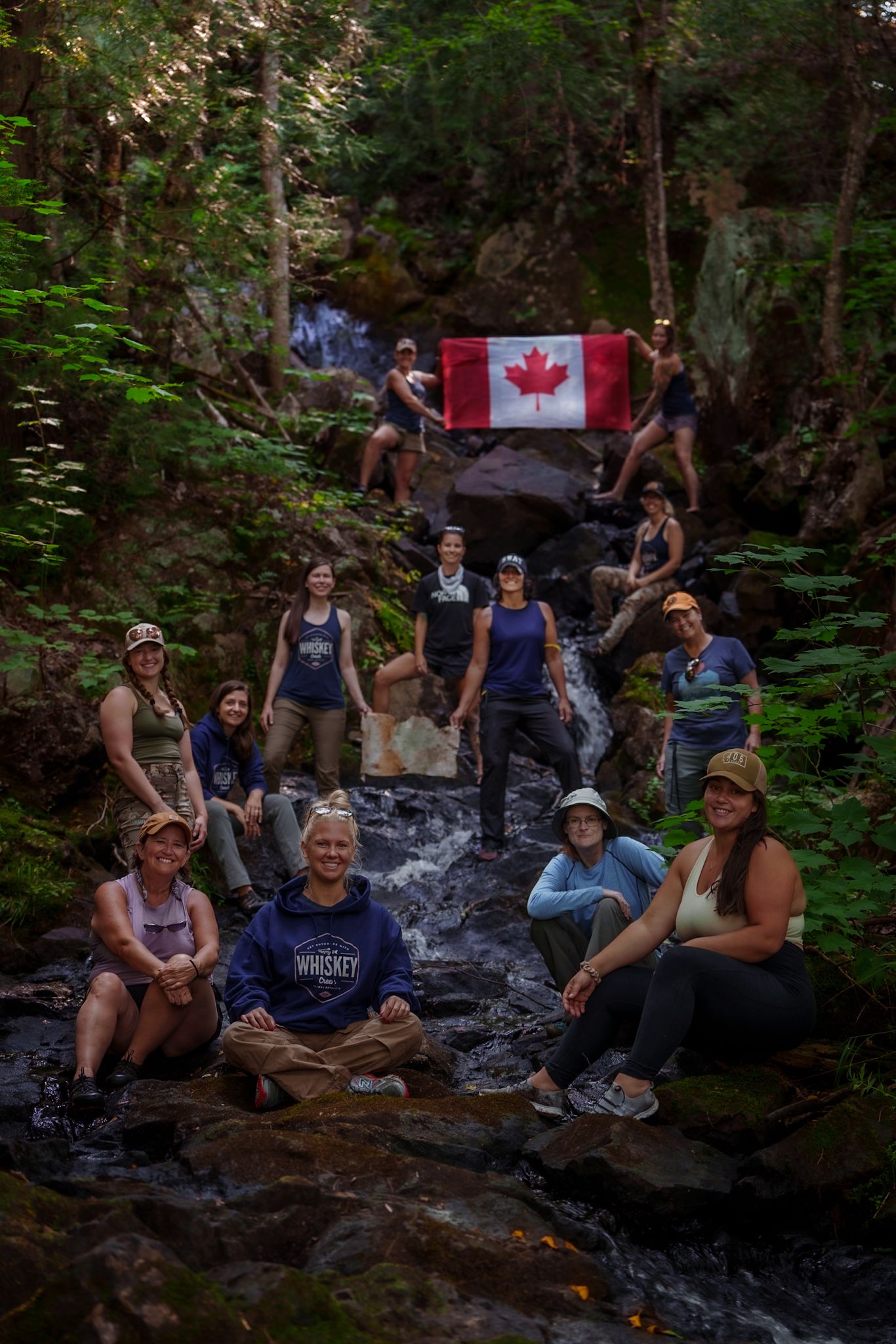A group of hikers pose with a Canadian flag at a cascading waterfall in a lush forest setting.