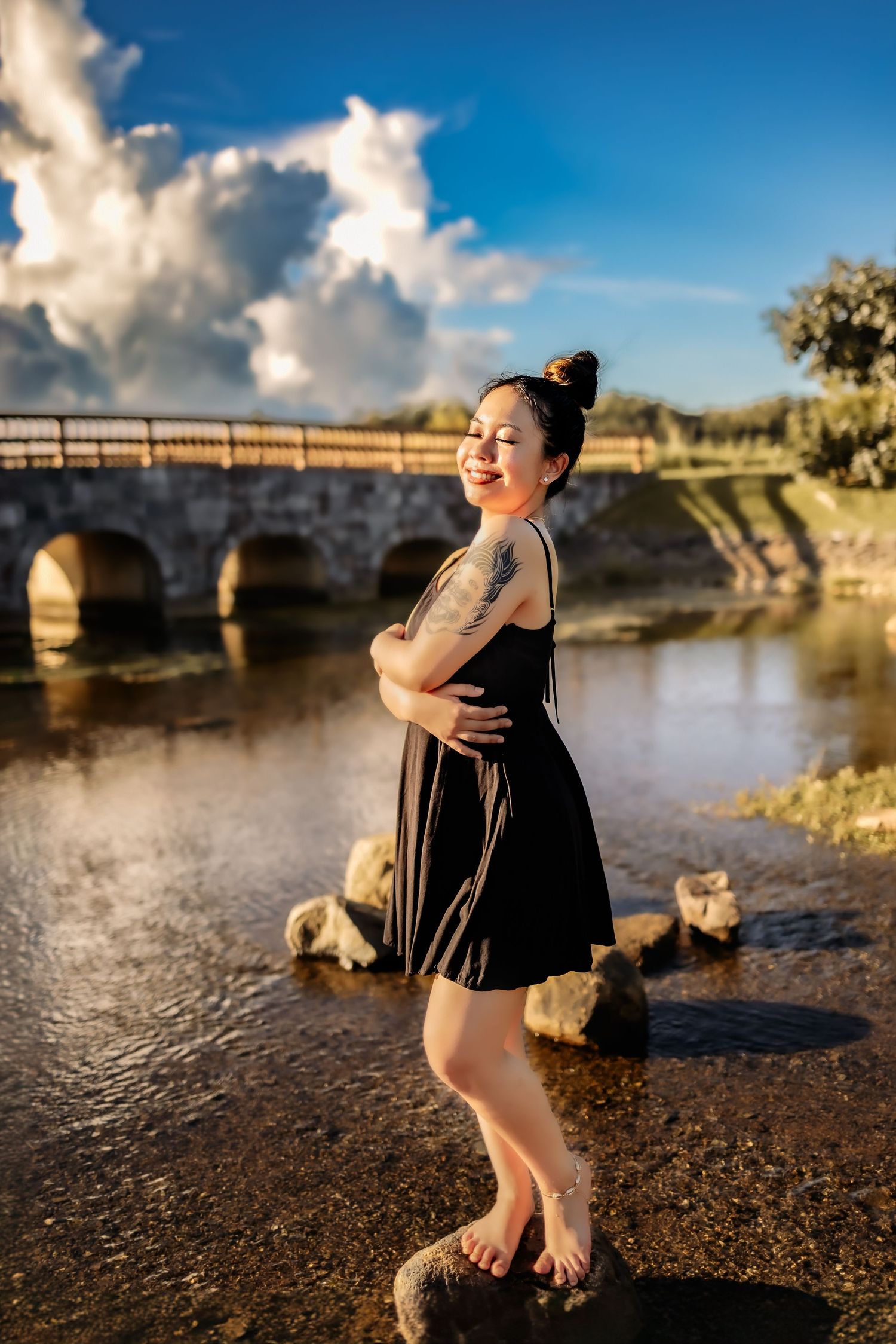 Person in black dress poses confidently by a stone bridge over calm water at sunset.