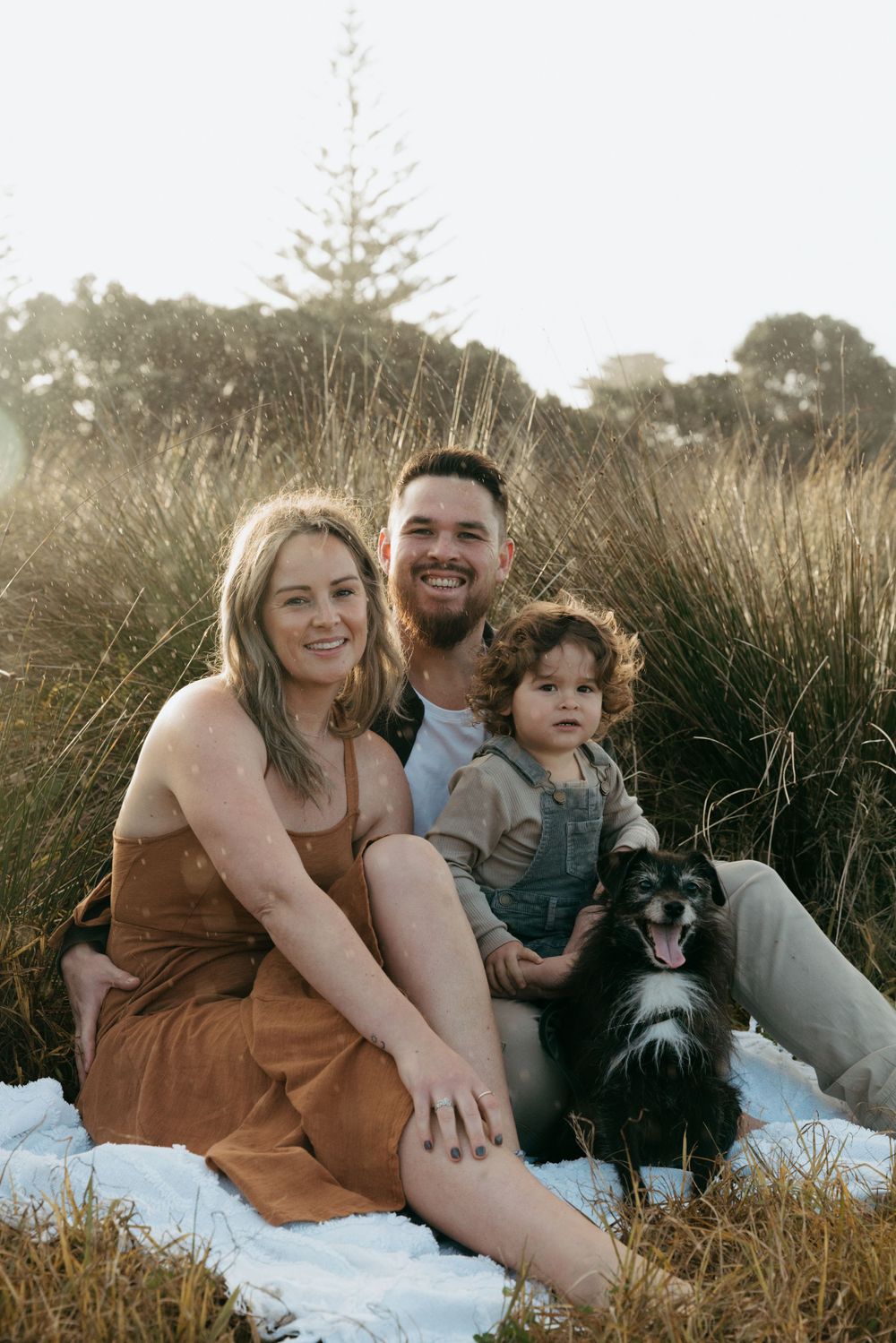 A family and their dog enjoy a picnic on a blanket in tall grass during golden hour.