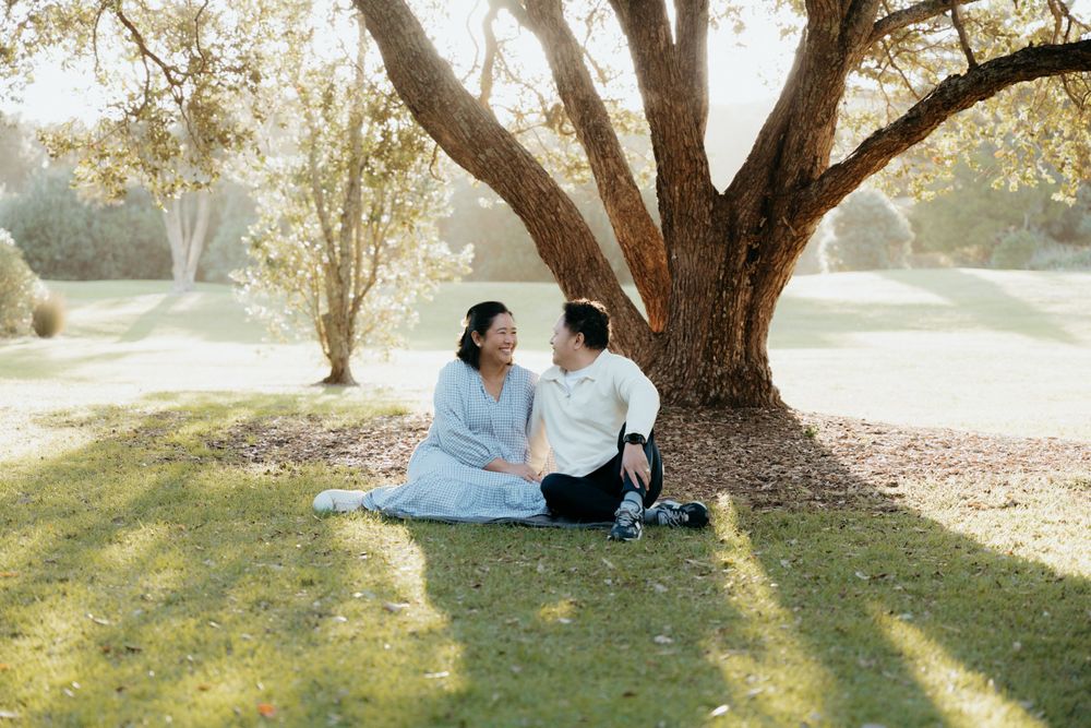 Couple sits together on blanket under large shady tree in peaceful park setting during golden hour.