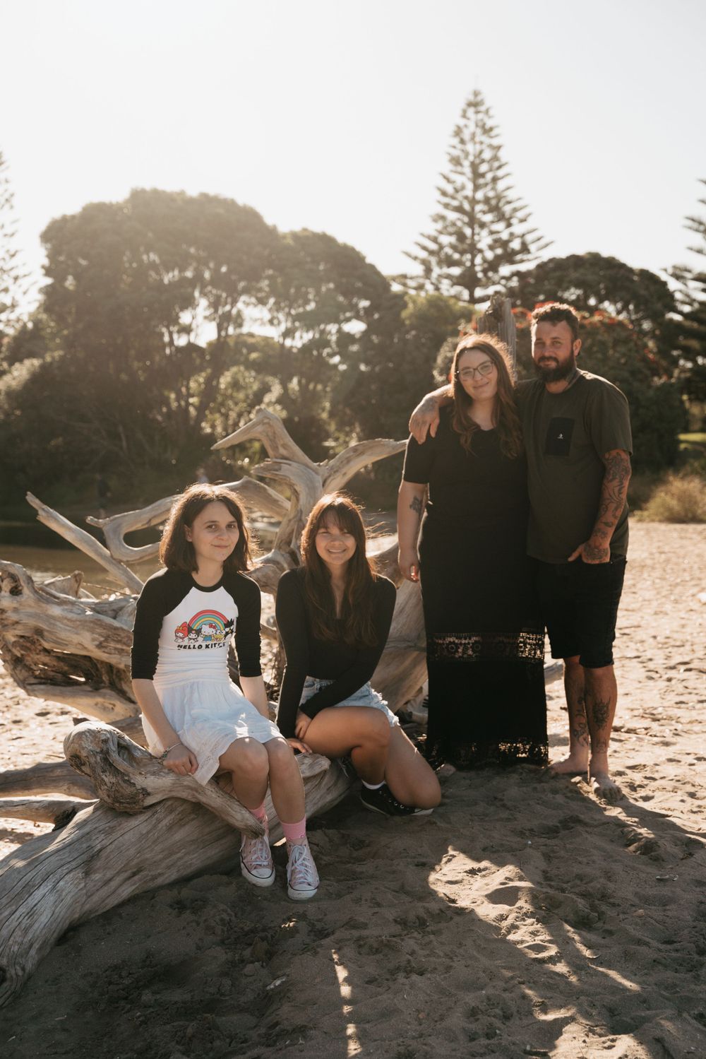 Group poses casually on driftwood at beach with pine trees in background during sunset.