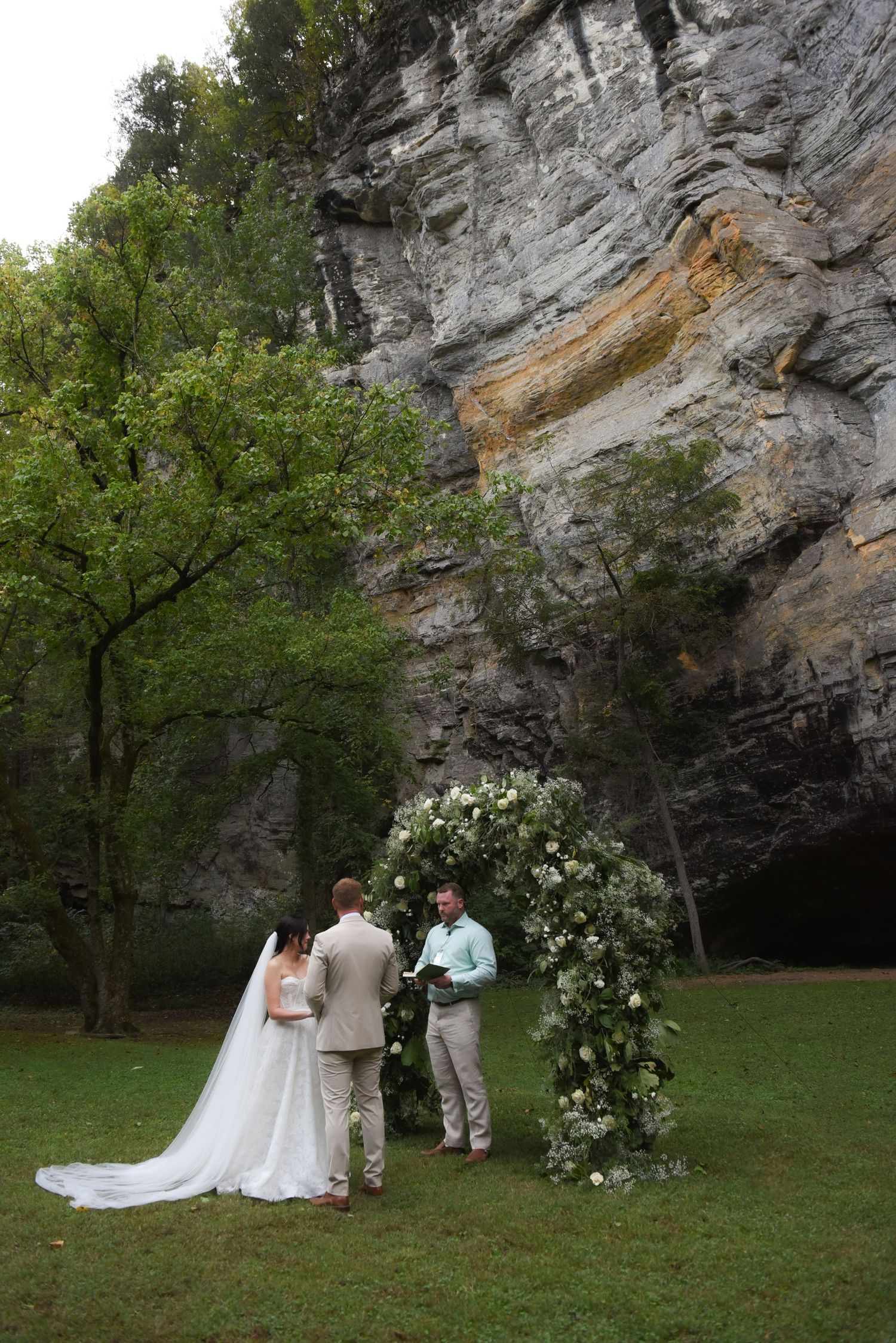 A wedding ceremony takes place in front of a majestic stone cliff face with floral decorations and greenery.