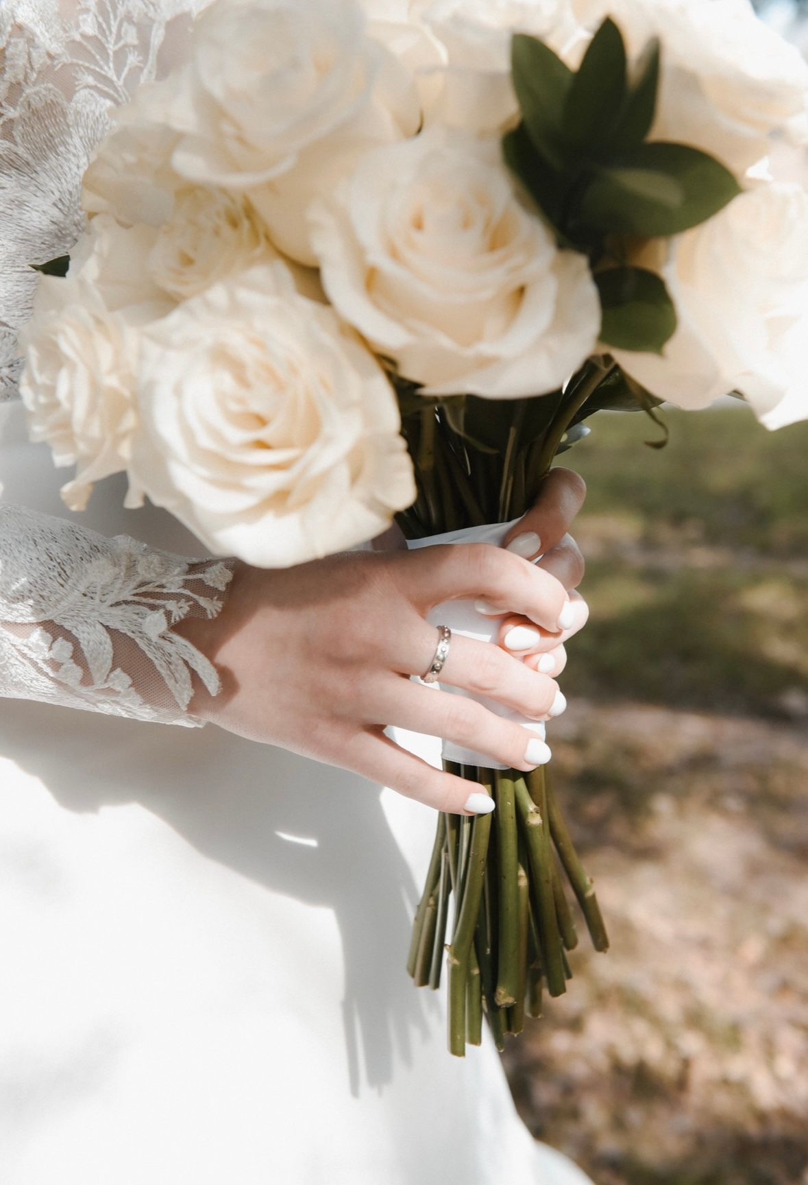 Classic white rose bridal bouquet held by hands wearing a lace sleeve and engagement ring in soft natural light.